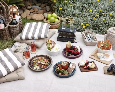 Picnic setup with striped cushions, assorted dishes, fruits, drinks, and decor; set on a blanket in a garden.