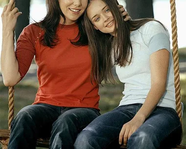 Two women from "Gilmore Girls" sit on a swing outdoors, smiling and leaning close together.