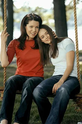 Two women from "Gilmore Girls" sit on a swing outdoors, smiling and leaning close together.