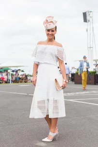 Woman in a white off-shoulder dress and pink floral hat at an outdoor event, holding a beige clutch.