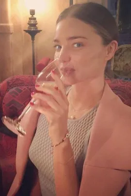 A woman in a grey dress, seated on a patterned chair, sips red wine in a cozy, warmly lit room.