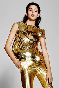 Woman in metallic gold top and pants posing with a neutral expression and one hand on hip.