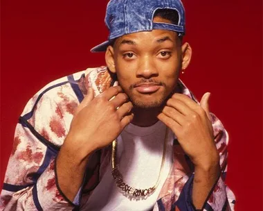 A young man wearing a colorful jacket, white shirt, denim cap, and gold necklace, posing against a red background.