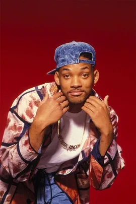 A young man wearing a colorful jacket, white shirt, denim cap, and gold necklace, posing against a red background.