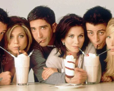 The six main characters from Friends sipping milkshakes, all leaning in towards the counter with playful expressions.