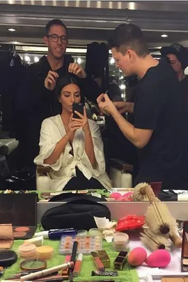 Woman in makeup chair using phone, makeup artists working on her hair and makeup, cosmetics scattered on table.