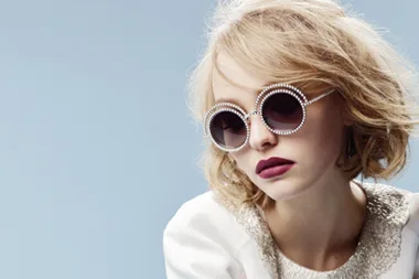 Woman with short blonde hair wearing round, pearl-studded sunglasses and white top against a blue background.