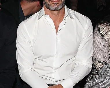 Marc Jacobs sitting at an event, wearing a white shirt and black pants, with folded hands on his lap.