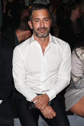 Marc Jacobs sitting at an event, wearing a white shirt and black pants, with folded hands on his lap.