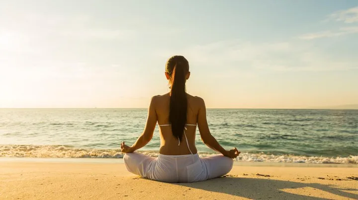 A person sitting cross-legged on a beach, meditating while facing the ocean at sunset.