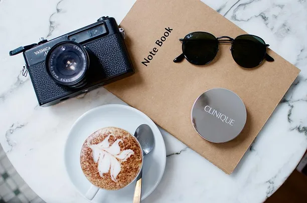 Camera, notebook, sunglasses, Clinique compact, and cappuccino on a marble table.