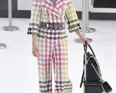 "Model in colorful checkered suit with rolling suitcase on Chanel runway at fashion show."