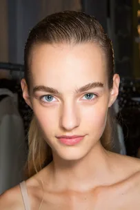 Young woman with slicked-back hair, natural makeup, and blue eyes, posing for a close-up photograph against a blurred background.