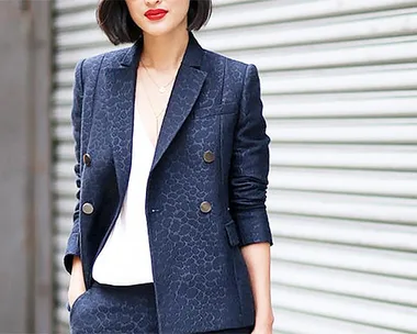 Woman in dark patterned suit with white blouse, standing against a roller door background.