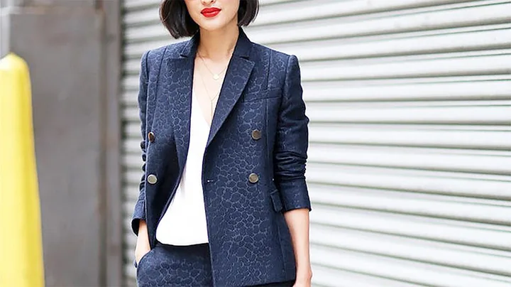 Woman in dark patterned suit with white blouse, standing against a roller door background.