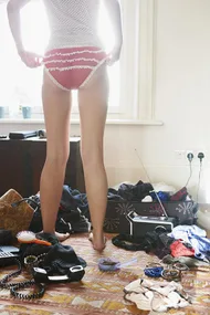 A person in red frilly underwear stands in a cluttered room, adjusting their waistband. Various items like clothes and accessories lie around.