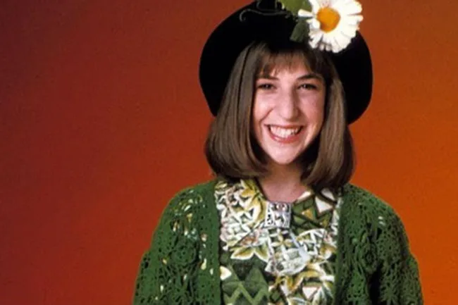 A person with a daisy hat smiles against an orange background, wearing a green crocheted sweater and patterned shirt.