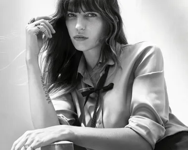 Black-and-white image of a woman with long hair and bangs, wearing a blouse with a ribbon tie, resting her chin on her hand.