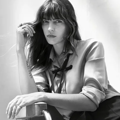 Black-and-white image of a woman with long hair and bangs, wearing a blouse with a ribbon tie, resting her chin on her hand.