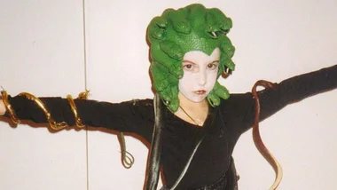 A young person dressed as Medusa with a green snake headdress, white face paint, and black outfit. Arms outstretched with toy snakes.