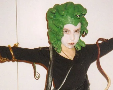 A young person dressed as Medusa with a green snake headdress, white face paint, and black outfit. Arms outstretched with toy snakes.