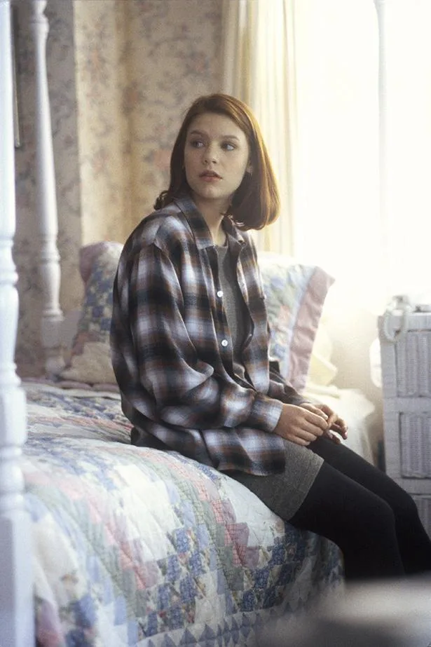 A young woman with short brown hair sits on a bed, wearing a plaid shirt, in a softly lit room.
