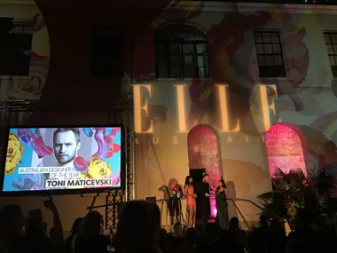 Projected image of Toni Maticevski at the Elle Style Awards 2015 with text "Australian Designer of the Year" and people on stage.