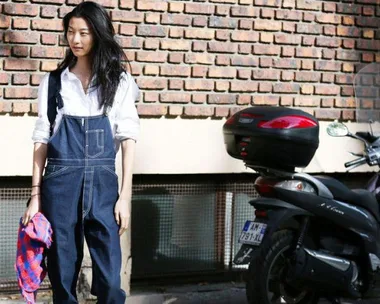 Woman in denim overalls stands beside a scooter, holding a red scarf, in front of a brick wall.