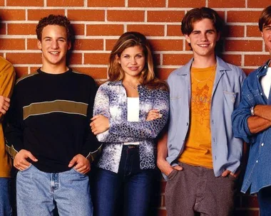 Cast of "Boy Meets World" in casual 90s clothing against a brick wall, including main characters Cory and Topanga.