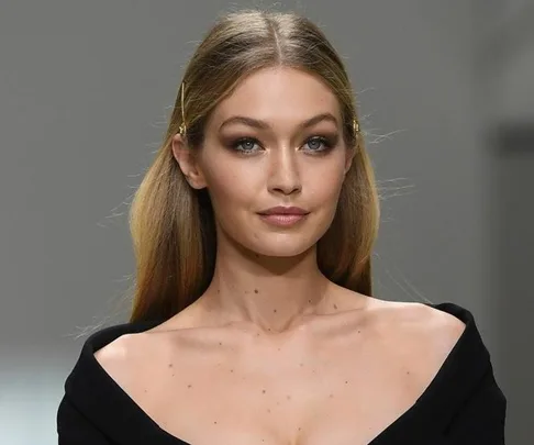 A woman with long hair, wearing a black top, displays a subtle nude lipstick shade on a runway.