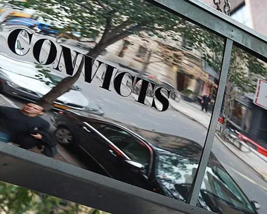 Hanging sign with "CONVICTS" reflected, in front of a street with parked cars and buildings in New York.