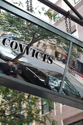 Hanging sign with "CONVICTS" reflected, in front of a street with parked cars and buildings in New York.