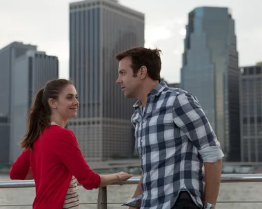 Alison Brie and Jason Sudeikis in a scene from the film "Sleeping with Other People," standing by a waterfront with city skyscrapers.