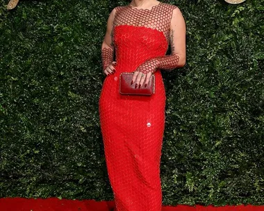 A person in a red sequined gown poses on a red carpet with a foliage backdrop at the British Fashion Awards.