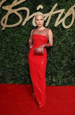 A person in a red sequined gown poses on a red carpet with a foliage backdrop at the British Fashion Awards.