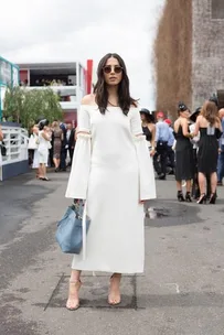 Woman in a white off-shoulder dress with bell sleeves, holding a blue bag, at Melbourne Spring Racing Carnival event.