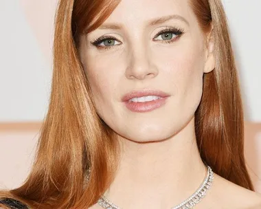 A woman with red hair, wearing a dark dress and a diamond necklace, poses for a photo against a light background.