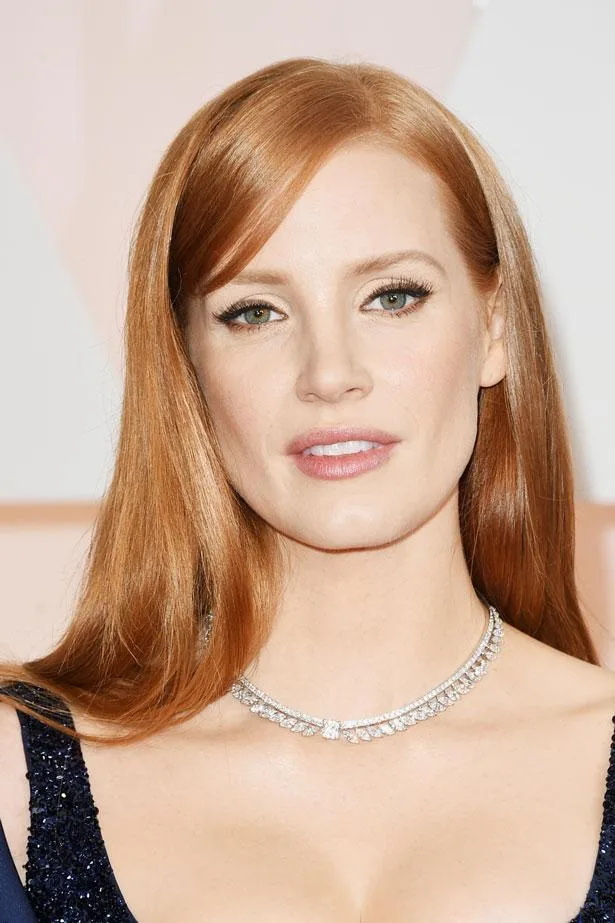 A woman with red hair, wearing a dark dress and a diamond necklace, poses for a photo against a light background.