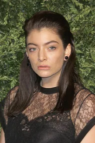 A person with straightened hair, wearing a lace top, poses in front of a green leafy background.