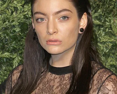 A person with straightened hair, wearing a lace top, poses in front of a green leafy background.