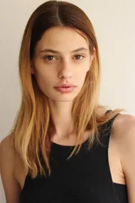 A female model with long straight hair, wearing a black sleeveless top, standing against a white wall.