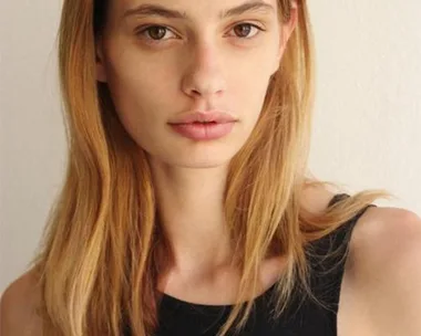 A female model with long straight hair, wearing a black sleeveless top, standing against a white wall.