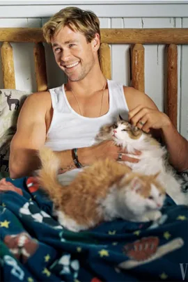 Chris Hemsworth in a white tank top cuddling with two kittens on a colorful blanket