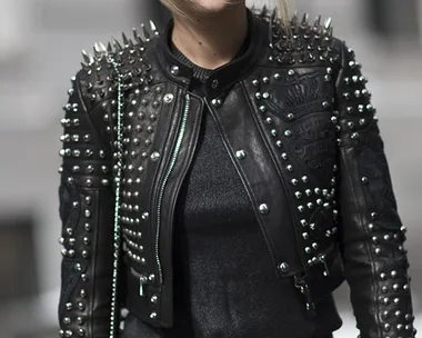 Woman in studded leather jacket, sunglasses, standing outdoors with gray background.