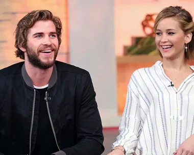 Actors, Liam Hemsworth and Jennifer Lawrence, smiling and seated during an interview on a TV show set.