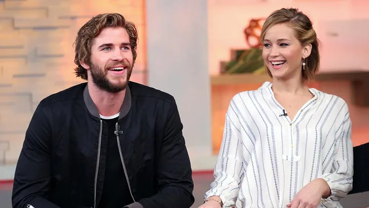 Actors, Liam Hemsworth and Jennifer Lawrence, smiling and seated during an interview on a TV show set.