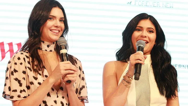 Two women with long dark hair hold microphones and smile on stage at an event.