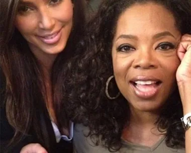 Two women smiling and posing for a photo, one with dark straight hair and the other with curly hair, in a warmly lit setting.