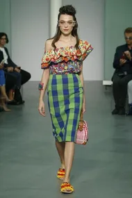 Model walking runway in colorful off-shoulder top, blue-green checked skirt, holding checkered handbag; seated audience in background.