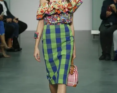 Model walking runway in colorful off-shoulder top, blue-green checked skirt, holding checkered handbag; seated audience in background.